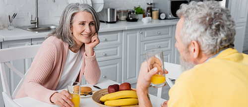 Middle aged woman holding glass of orange juice near breakfast and blurred husband, banner