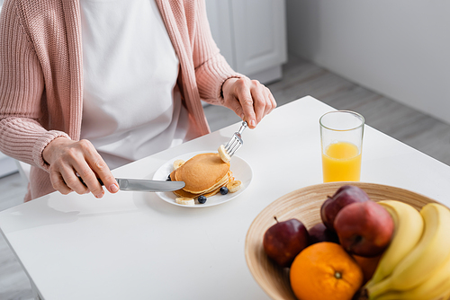 Cropped view of mature woman holding cutlery near pancakes and fruits at home