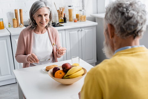 Cheerful mature woman looking at blurred husband near pancakes and fruits in kitchen