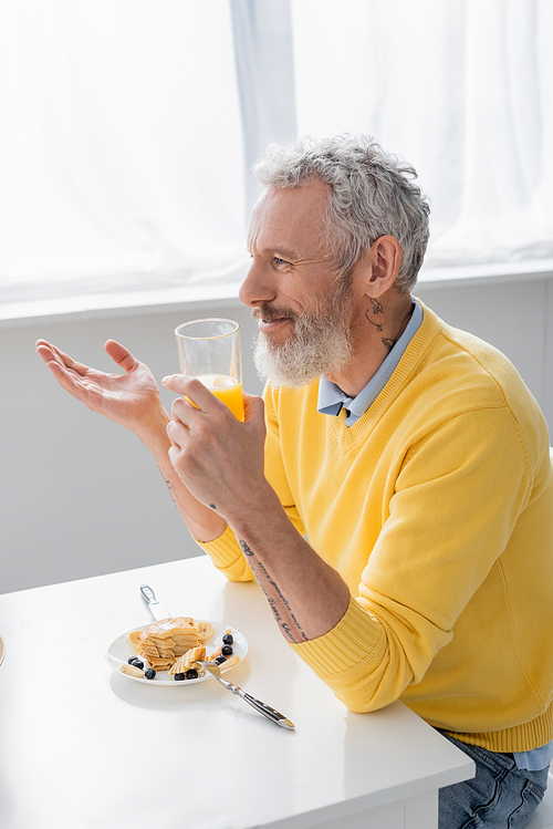 Smiling mature man holding orange juice near pancakes in kitchen