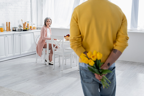 Smiling woman looking at blurred husband hiding flowers in kitchen