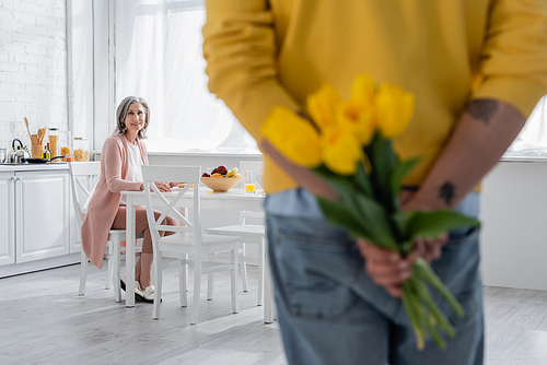 Middle aged woman looking at husband hiding tulips in kitchen
