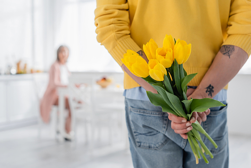 Mature man hiding flowers near blurred woman in kitchen