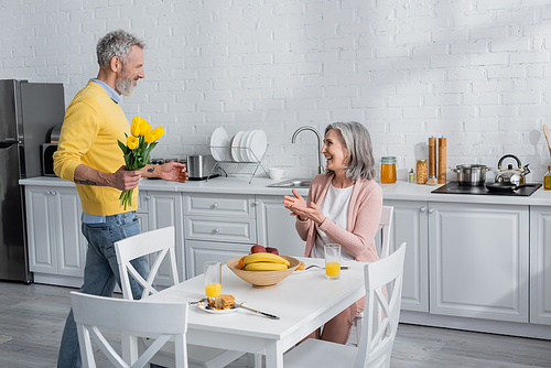 Side view of man holding flowers near positive wife and breakfast in kitchen