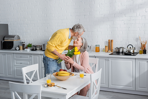 Man with flowers kissing wife during breakfast in kitchen
