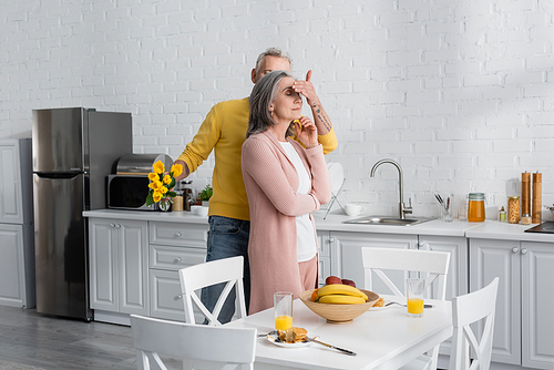 Man holding flowers and covering eyes of wife near breakfast in kitchen