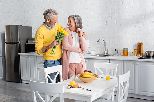 Cheerful man holding tulips near mature wife and tasty breakfast in kitchen