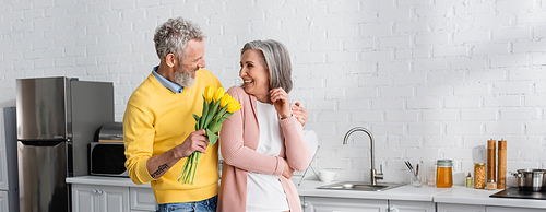 Smiling mature man hugging wife and holding yellow tulips in kitchen, banner
