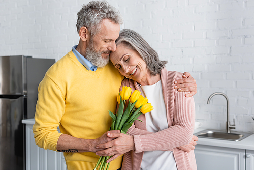 Middle aged man holding tulips near wife with closed eyes in kitchen