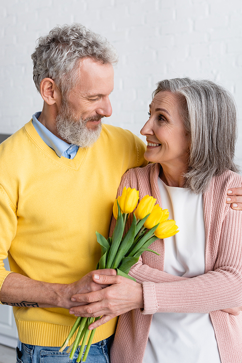 Smiling woman holding flowers near bearded husband at home