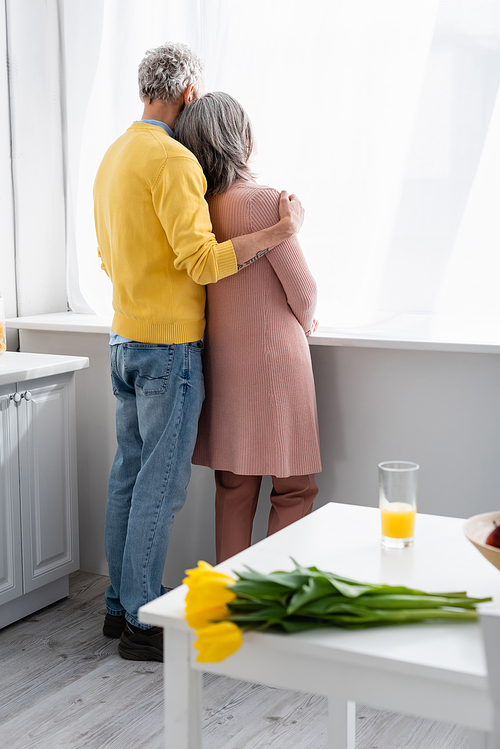 Back view of mature couple hugging near window and blurred tulips on table at home