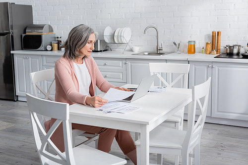 Middle aged woman using laptop near papers with bills in kitchen
