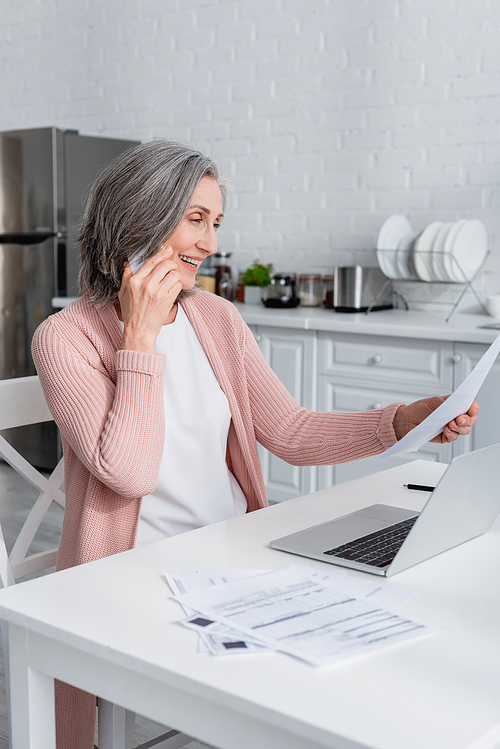Smiling mature woman talking on smartphone and holding paper near laptop in kitchen