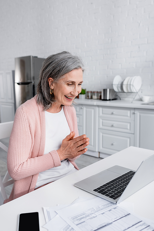 Grey haired woman smiling and doing pray gesture near devices and bills in kitchen