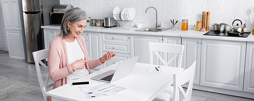 Mature woman gesturing during video call on laptop near smartphone and bills in kitchen, banner