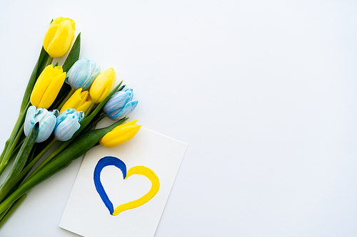 Top view of card with pained heart symbol near yellow and blue tulips on white background