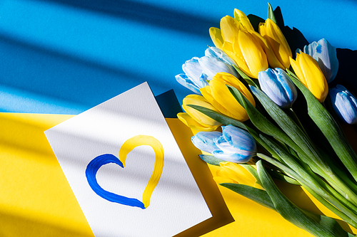 Top view of card with painted heart sign near blue and yellow tulips on ukrainian flag