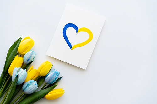 Top view of card with painted blue and yellow heart symbol near flowers on white background