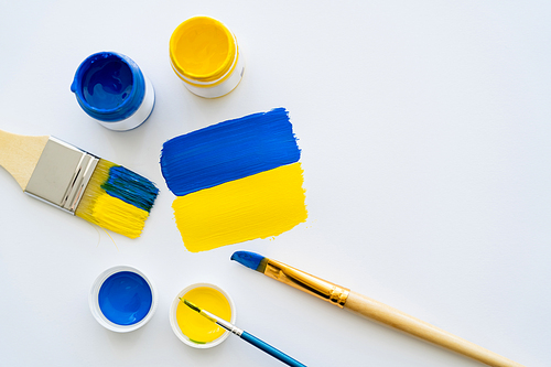 Top view of paints and paintbrushes near painted ukrainian flag on white background