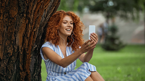 happy redhead woman in blue dress and earphones listening music while looking at smartphone