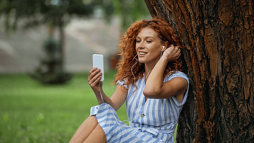 pleased redhead woman in blue dress and earphones listening music while looking at smartphone
