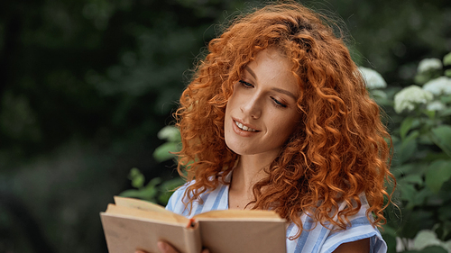 curly redhead woman smiling while reading book outdoors