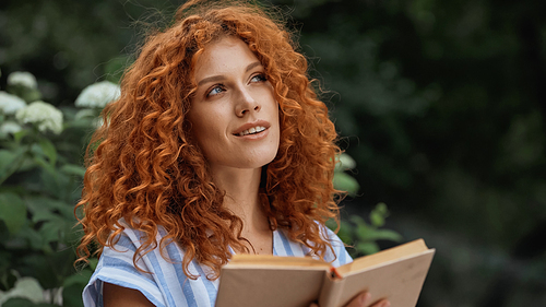 dreamy redhead woman smiling while holding book outdoors