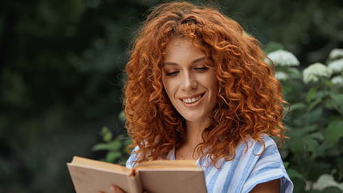 happy redhead woman smiling while reading book outdoors
