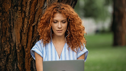 young redhead woman listening music in earphones while using laptop in park
