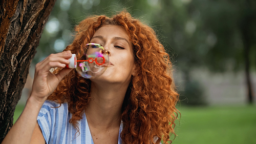 curly redhead woman blowing soap bubble in park