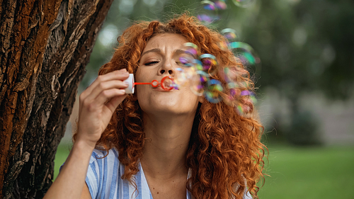curly redhead woman frowning while blowing soap bubbles in park