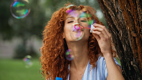 cheerful redhead woman blowing soap bubbles in park