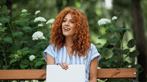 happy redhead woman using laptop while sitting on bench in park