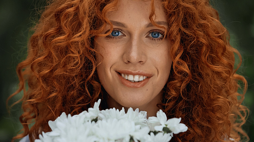 close up of curly redhead woman with blue eyes smiling near white flowers