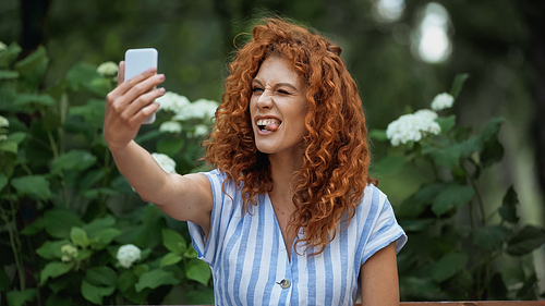 funny redhead woman sticking out tongue while taking selfie in green park