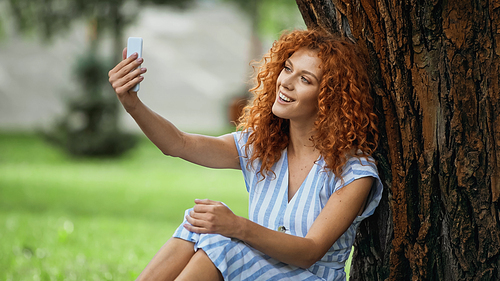 happy redhead woman taking selfie while sitting under tree trunk