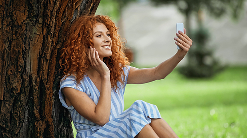 pleased redhead woman taking selfie while sitting under tree trunk