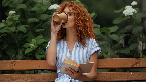 curly redhead woman in striped dress holding book and drinking coffee to go while sitting on bench in park