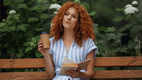 pleased redhead woman in striped dress holding book and paper cup while sitting on bench in park