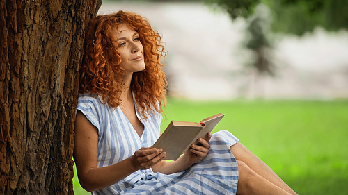pleased redhead woman in striped dress holding book and sitting under tree trunk