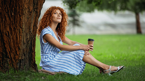 dreamy redhead woman in blue dress sitting under tree trunk and holding coffee to go