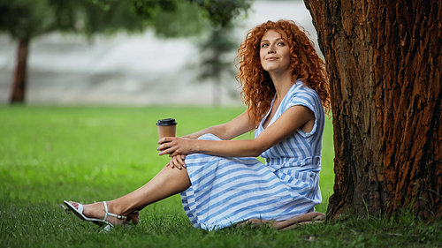 pleased redhead woman in blue dress sitting under tree trunk and holding coffee to go