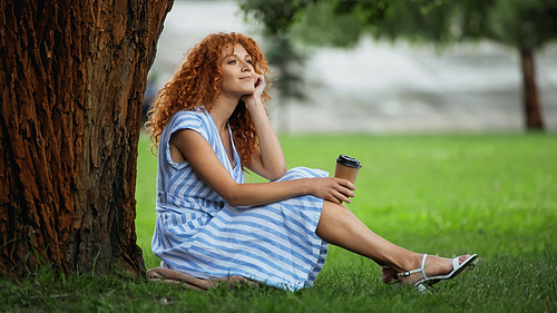 dreamy redhead woman in blue dress smiling while sitting under tree trunk and holding coffee to go