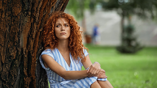 upset redhead woman in blue dress sitting under tree trunk in park