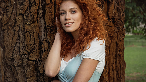 young and curly redhead woman smiling near tree in park