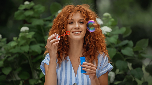 cheerful redhead woman looking at blurred soap bubbles in park