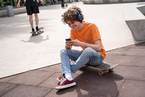 happy man in headphones sitting on skateboard and using smartphone in skate park
