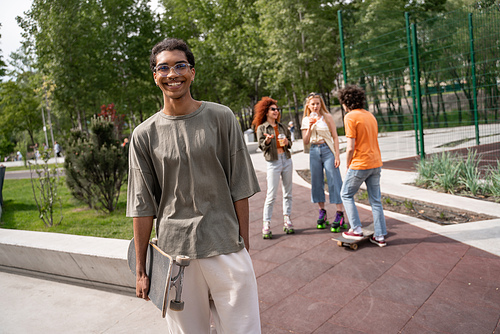 young and cheerful african american man holding skate near friends on background
