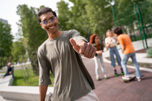 blurred african american man showing thumb up near blurred skaters in park