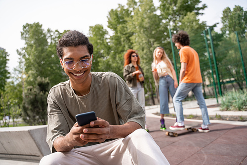 african american man in eyeglasses messaging on cellphone near skaters on blurred background